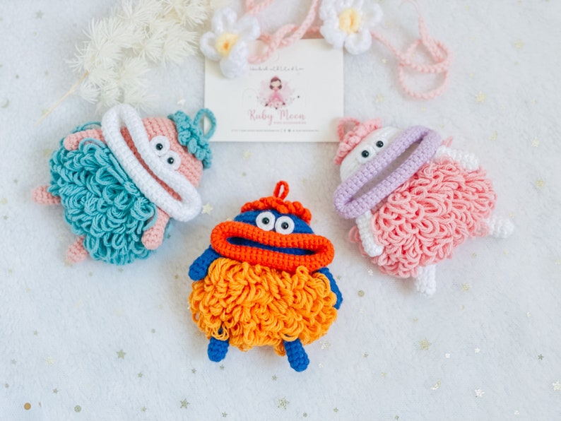 Puede incluir: Tres juguetes de peluche tejidos a crochet con diferentes colores de pelo y rasgos faciales. Los juguetes son azul, rosa y naranja. El juguete azul tiene una boca roja y pelo naranja. El juguete rosa tiene una boca morada y pelo rosa. El juguete naranja tiene una boca azul y pelo naranja.
