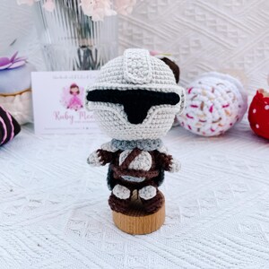 May include: A handmade crochet figure of a character from a science fiction franchise. The figure is primarily light gray with black accents, brown details, and a wooden base. The figure is standing on a wooden base. The figure is handmade.