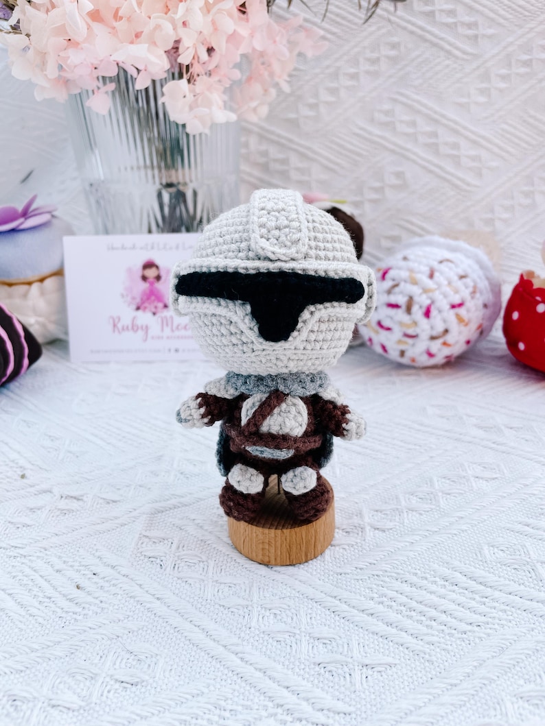May include: A handmade crochet figure of a character from a science fiction franchise. The figure is primarily light gray with black accents, brown details, and a wooden base. The figure is standing on a wooden base. The figure is handmade.