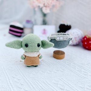 May include: A handmade crochet figure of a character with large ears, green skin, and a tan outfit. The figure is standing on a white surface. A small gray crochet basket with white buttons is nearby. Other decorative items are in the background.