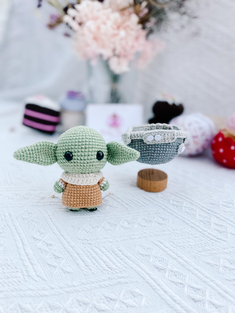May include: A handmade crochet figure of a character with large ears, green skin, and a tan outfit. The figure is standing on a white surface. A small gray crochet basket with white buttons is nearby. Other decorative items are in the background.