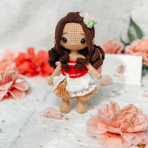 May include: A handmade crochet doll with brown hair, wearing a white and red dress with a tan skirt. The doll has a flower in its hair and is surrounded by pink flowers. The doll is a handmade craft.