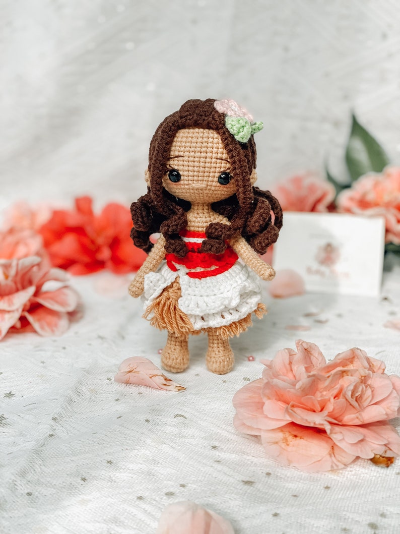 May include: A handmade crochet doll with brown hair, wearing a white and red dress with a tan skirt. The doll has a flower in its hair and is surrounded by pink flowers. The doll is a handmade craft.