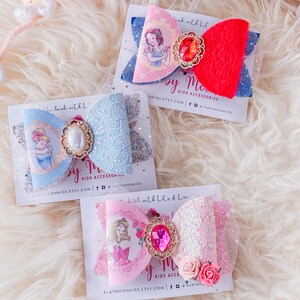 May include: Three handmade hair bows featuring Disney princesses Snow White, Cinderella, and Aurora. Each bow is made with a different color of tulle and lace, and features a jeweled center piece. The bows are displayed on white cards with the text "Made with Lots of Love" and the Etsy shop name "Ruby Moon Kids".