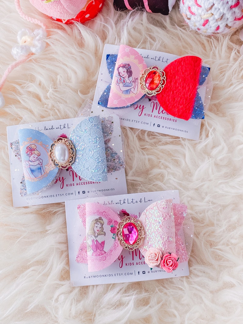 May include: Three handmade hair bows featuring Disney princesses Snow White, Cinderella, and Aurora. Each bow is made with a different color of tulle and lace, and features a jeweled center piece. The bows are displayed on white cards with the text "Made with Lots of Love" and the Etsy shop name "Ruby Moon Kids".