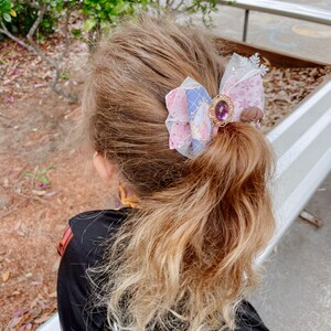 May include: A young girl with long, wavy, light brown hair with blonde highlights wears a black dress and has a pink, purple, and blue bow with a jeweled center in her hair.