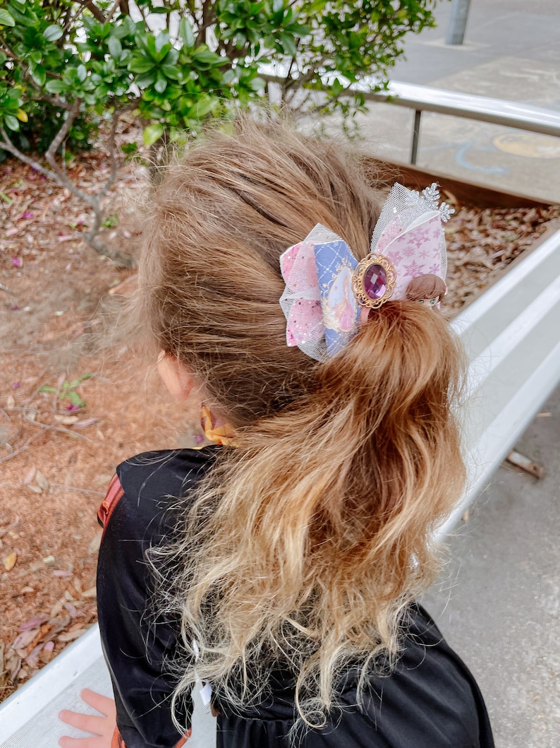 May include: A young girl with long, wavy, light brown hair with blonde highlights wears a black dress and has a pink, purple, and blue bow with a jeweled center in her hair.