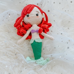May include: A crocheted mermaid doll with red hair, a green tail, and a purple seashell bra. The doll is sitting on a white background with silver glitter stars.