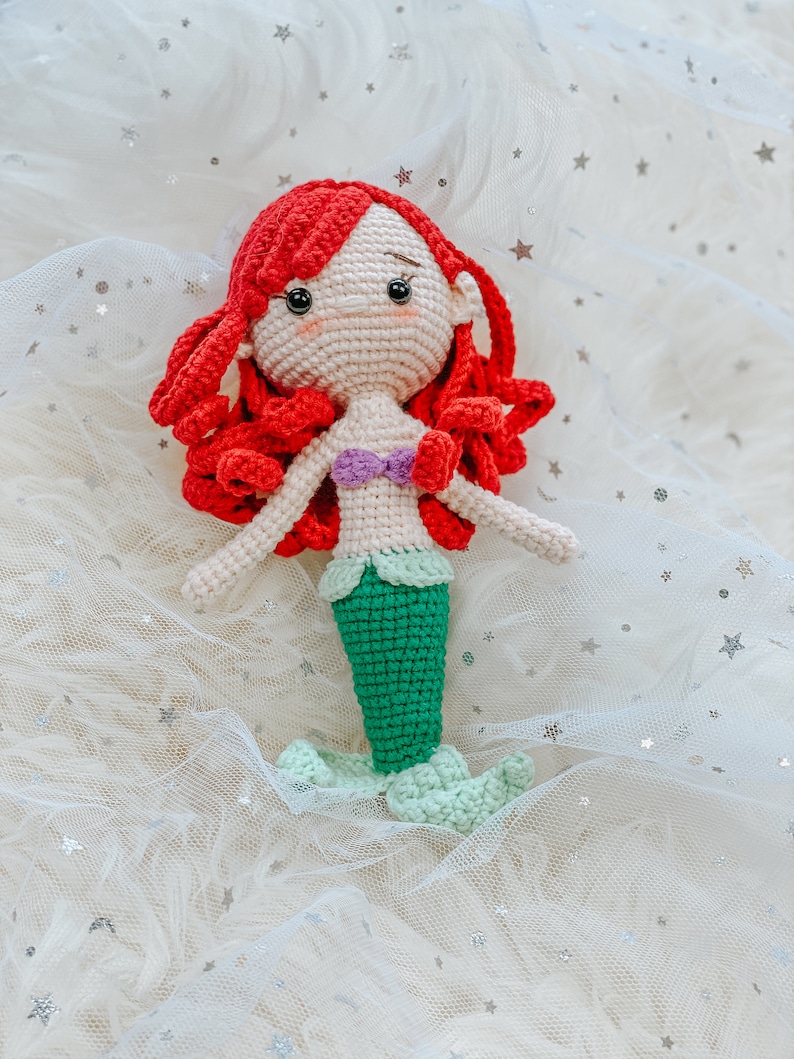 May include: A crocheted mermaid doll with red hair, a green tail, and a purple seashell bra. The doll is sitting on a white background with silver glitter stars.