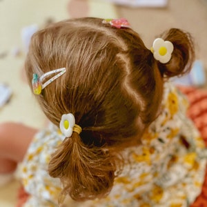 K&ouml;nnte beinhalten: Ein junges M&auml;dchen mit langen braunen Haaren, das ein gelbes und wei&szlig;es Blumenkleid tr&auml;gt. Sie hat zwei wei&szlig;e Blumenhaarklammern im Haar und eine rosa und wei&szlig;e Haarklammer.