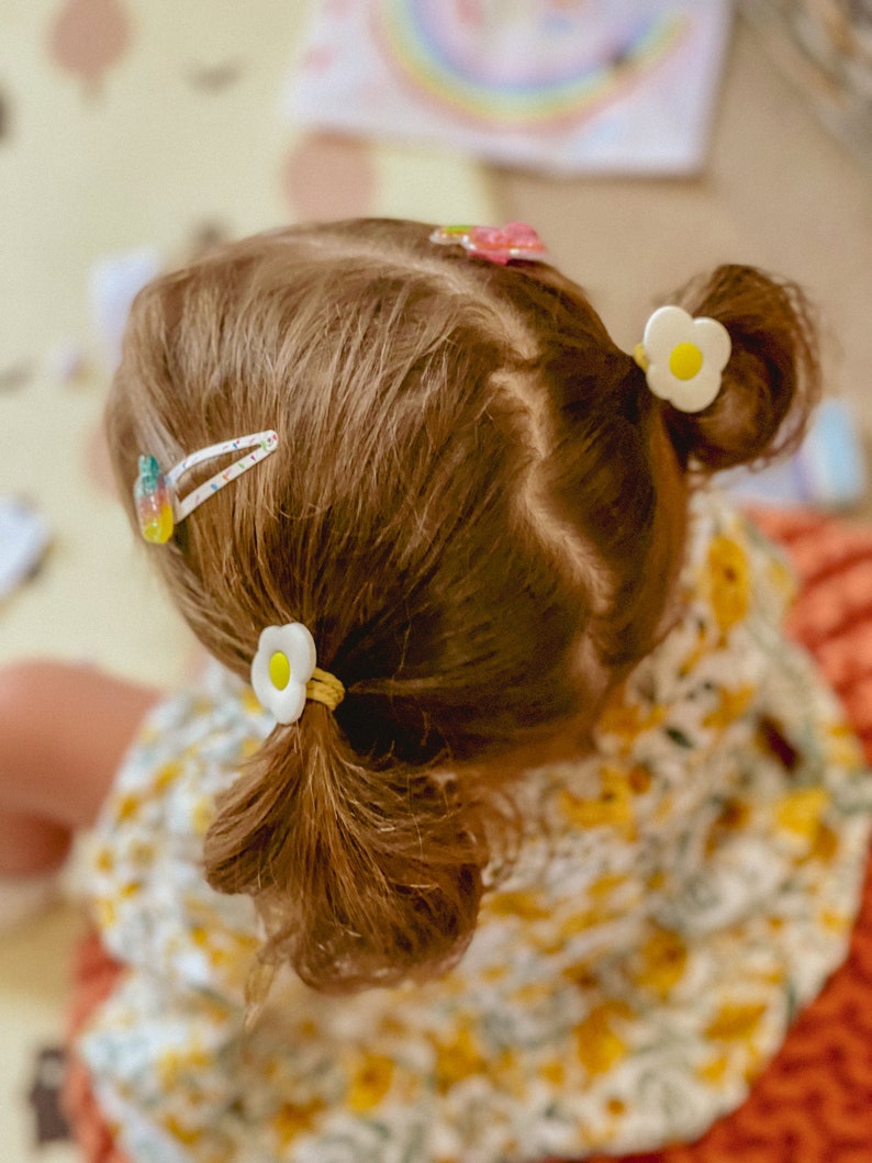 K&ouml;nnte beinhalten: Ein junges M&auml;dchen mit langen braunen Haaren, das ein gelbes und wei&szlig;es Blumenkleid tr&auml;gt. Sie hat zwei wei&szlig;e Blumenhaarklammern im Haar und eine rosa und wei&szlig;e Haarklammer.