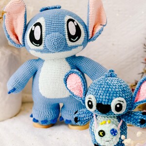 Op de afbeelding: Twee blauwe gehaakte Stitch knuffels. De grotere Stitch staat op een houten voet en heeft roze oren. De kleinere Stitch zit op een houten voet en heeft een groene gehaakte tas met een zwarte X.