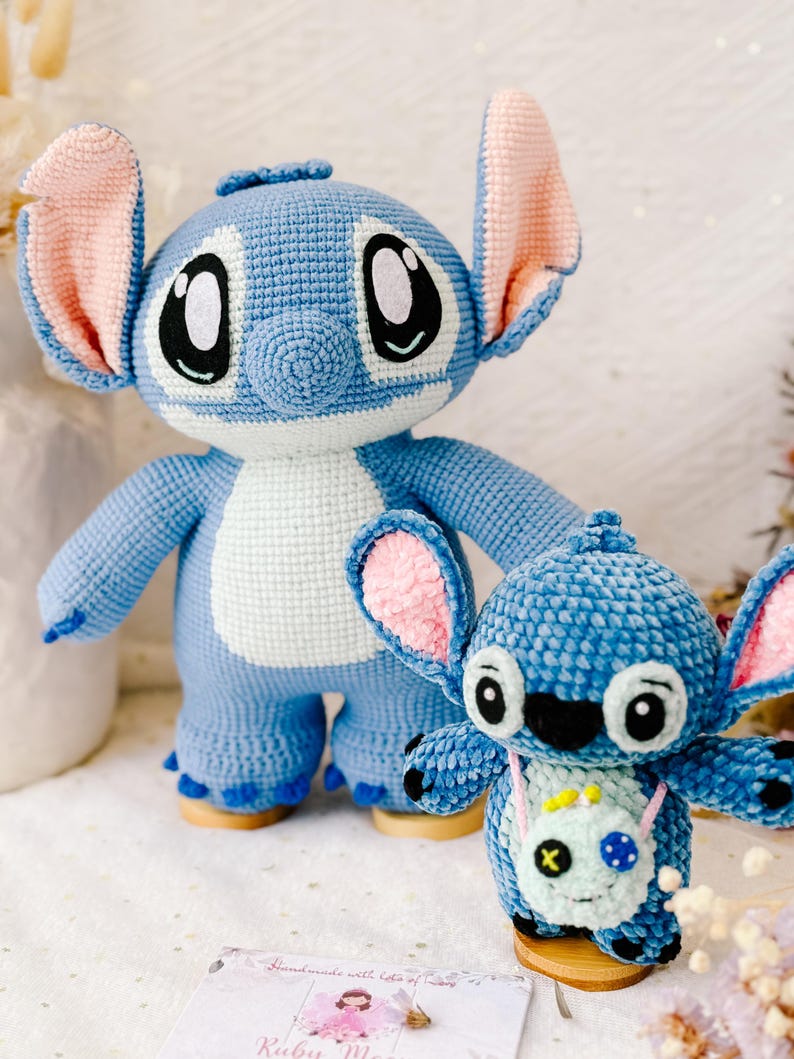 Op de afbeelding: Twee blauwe gehaakte Stitch knuffels. De grotere Stitch staat op een houten voet en heeft roze oren. De kleinere Stitch zit op een houten voet en heeft een groene gehaakte tas met een zwarte X.