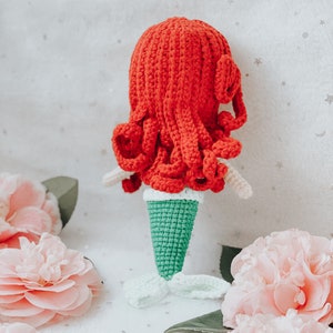 May include: A crocheted mermaid doll with red hair and a green tail. The doll is sitting on a white surface with pink flowers.