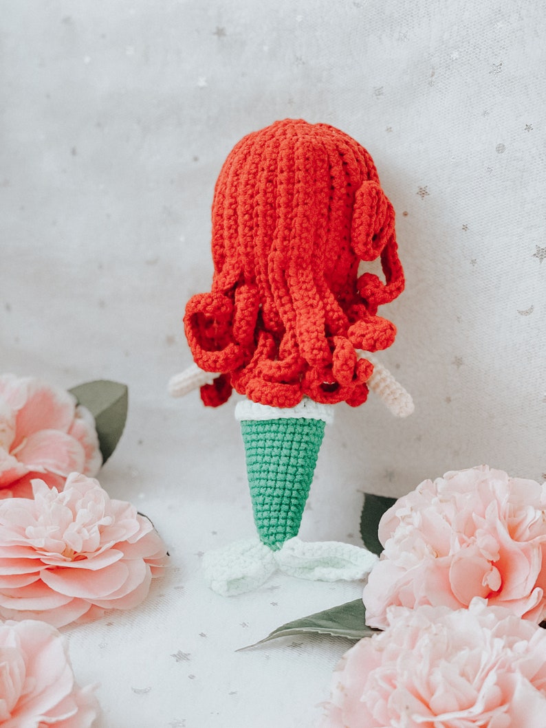 May include: A crocheted mermaid doll with red hair and a green tail. The doll is sitting on a white surface with pink flowers.