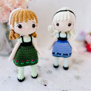Puede incluir: Dos mu&ntilde;ecas de crochet, una con cabello casta&ntilde;o y un vestido verde, la otra con cabello rubio y un vestido azul. Las mu&ntilde;ecas est&aacute;n de pie sobre una superficie blanca con copos de nieve plateados.