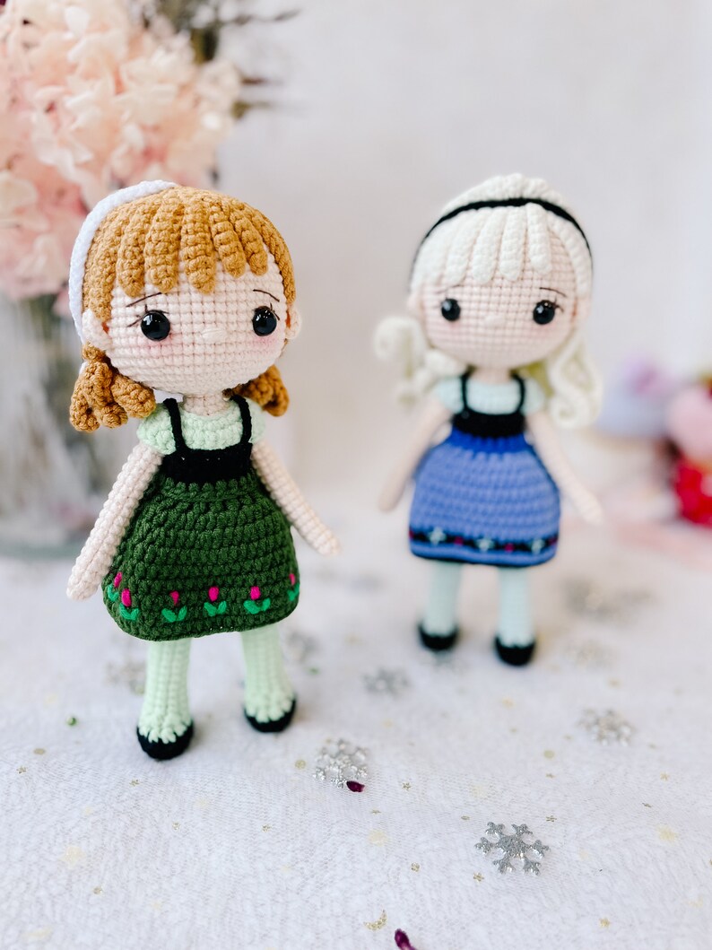 Puede incluir: Dos mu&ntilde;ecas de crochet, una con cabello casta&ntilde;o y un vestido verde, la otra con cabello rubio y un vestido azul. Las mu&ntilde;ecas est&aacute;n de pie sobre una superficie blanca con copos de nieve plateados.