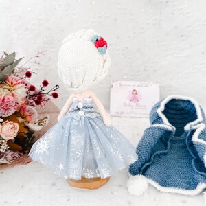 May include: A handmade crochet doll wearing a light blue dress with silver snowflake accents and a matching blue cape. The doll has a white crocheted hairstyle with a red and blue flower. A small business card is in the background.