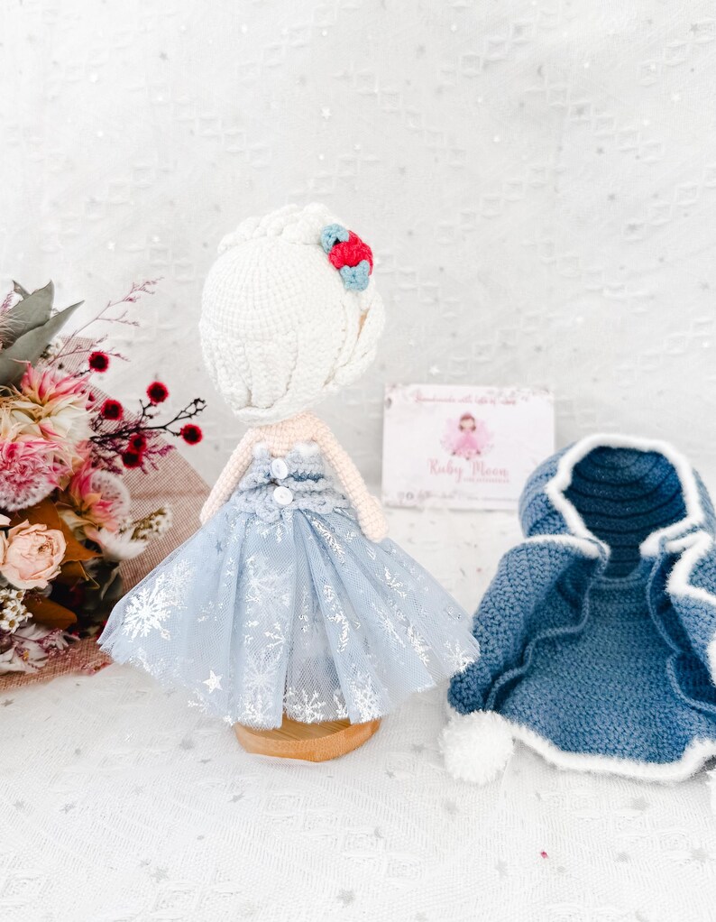 May include: A handmade crochet doll wearing a light blue dress with silver snowflake accents and a matching blue cape. The doll has a white crocheted hairstyle with a red and blue flower. A small business card is in the background.