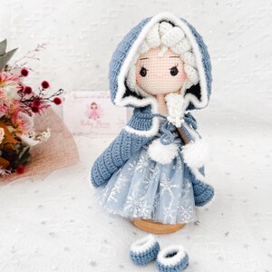 May include: A handmade crochet doll wearing a blue hooded cape with white trim and a snowflake-patterned dress. The doll has a light skin tone, rosy cheeks, and is accompanied by matching blue booties. The doll is a charming, handcrafted item.