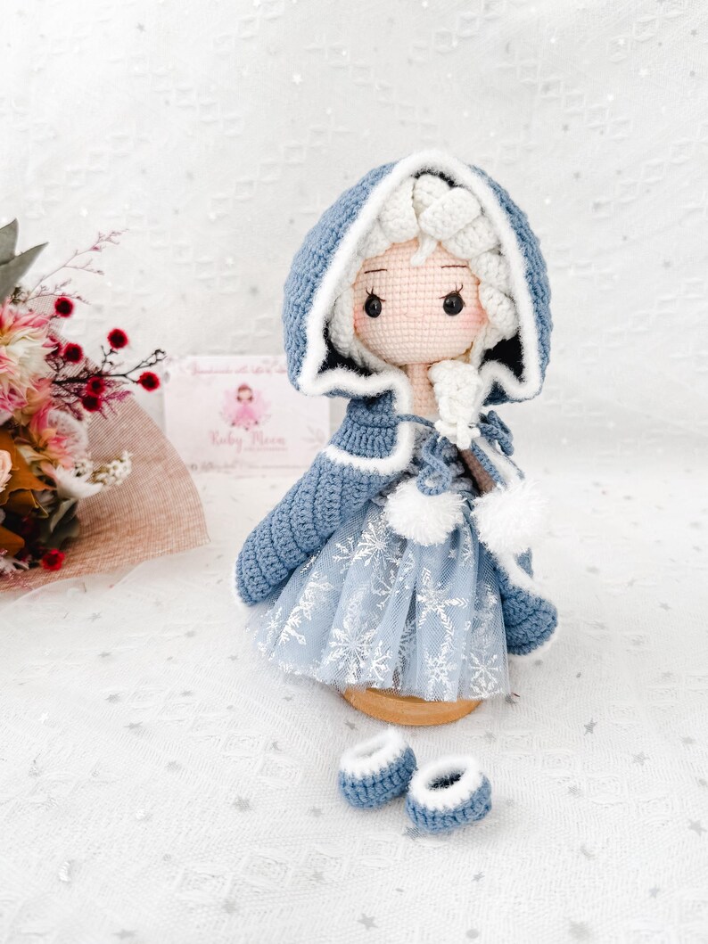 May include: A handmade crochet doll wearing a blue hooded cape with white trim and a snowflake-patterned dress. The doll has a light skin tone, rosy cheeks, and is accompanied by matching blue booties. The doll is a charming, handcrafted item.
