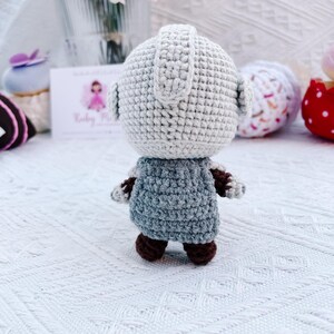 May include: A handmade crochet doll with a light gray head and a blue-gray body. The doll has brown feet and arms. The doll is standing on a white surface. The background includes flowers and other decorative items.