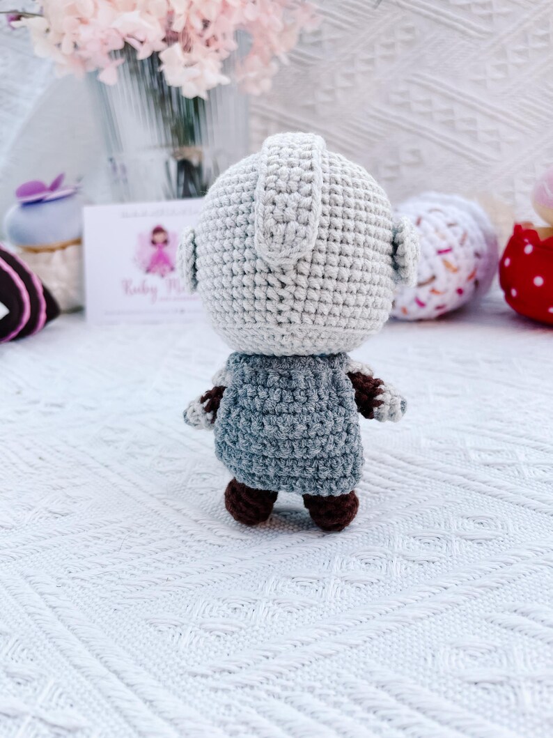 May include: A handmade crochet doll with a light gray head and a blue-gray body. The doll has brown feet and arms. The doll is standing on a white surface. The background includes flowers and other decorative items.