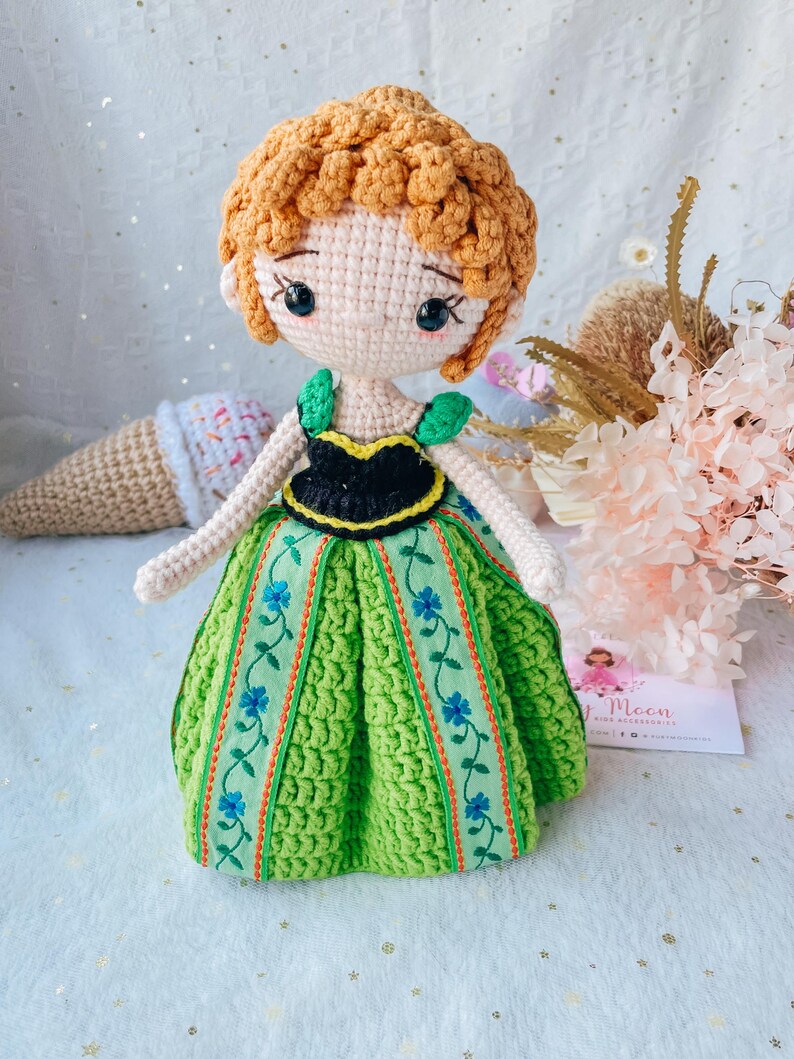 May include: Crocheted doll with brown hair, a green dress with floral embroidery, and a black bodice. The doll has a sweet, cartoon-like face with large eyes.