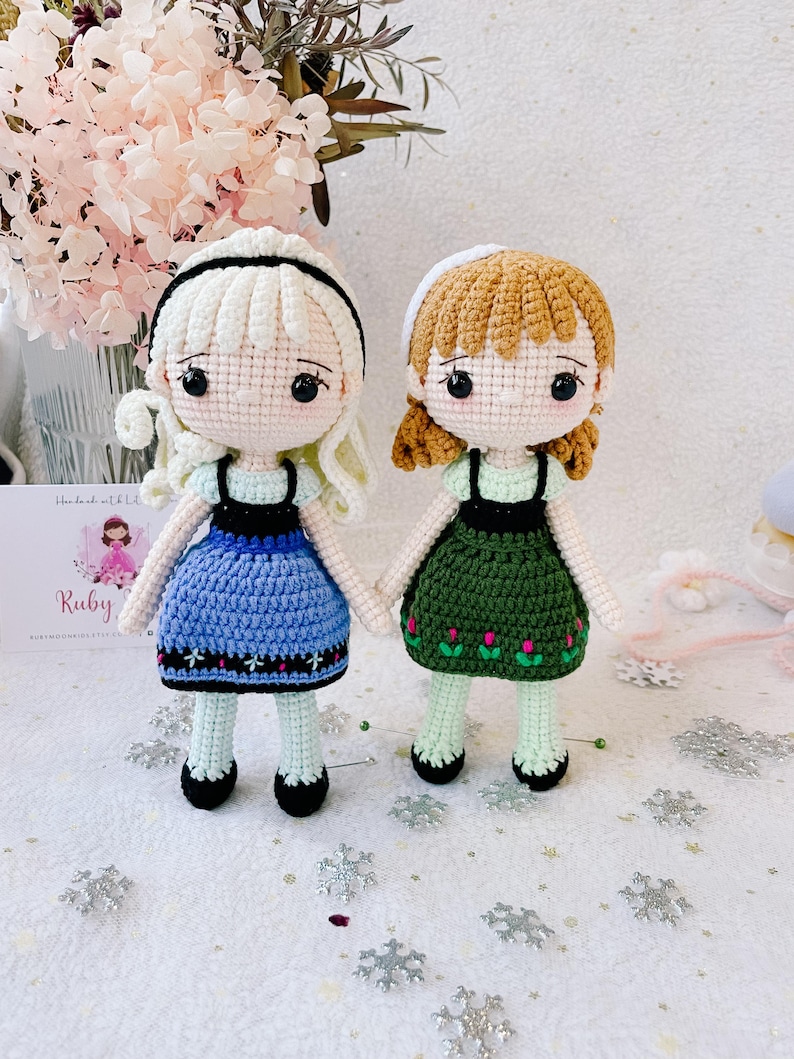 Puede incluir: Dos mu&ntilde;ecas de crochet, una con un vestido azul y la otra con un vestido verde, de pie sobre una superficie blanca con copos de nieve plateados. Las mu&ntilde;ecas tienen el pelo rubio y los ojos negros. Las mu&ntilde;ecas est&aacute;n hechas a mano y tienen el texto "Handmade with Love" y "Ruby" en una tarjeta en el fondo.