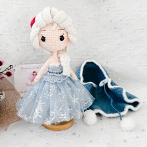 May include: A handmade crochet doll with white hair, wearing a light blue dress with snowflake accents. A matching blue cape with white trim and pom-poms sits nearby. The doll is on a wooden stand, showcasing a whimsical, handcrafted design.