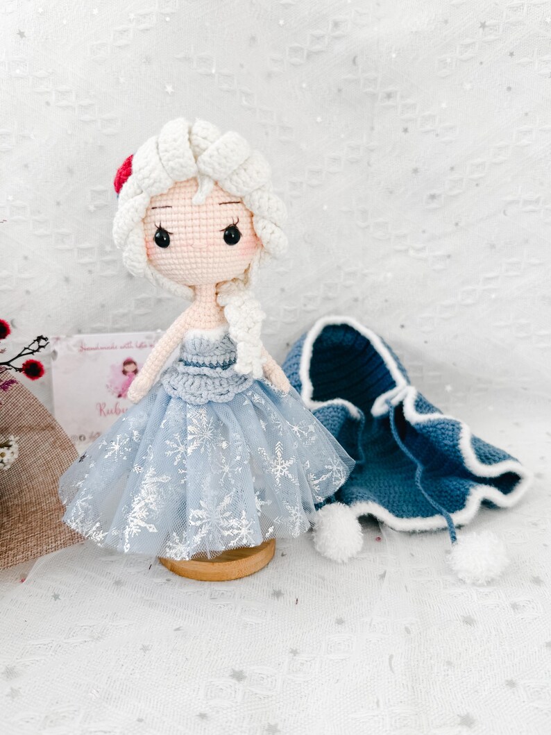 May include: A handmade crochet doll with white hair, wearing a light blue dress with snowflake accents. A matching blue cape with white trim and pom-poms sits nearby. The doll is on a wooden stand, showcasing a whimsical, handcrafted design.