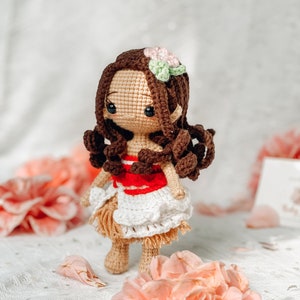 May include: A handmade crochet doll with brown hair, a flower hair accessory, and a tan skin tone. The doll wears a red and white top, a white skirt, and a tan skirt. The doll is surrounded by pink flowers.