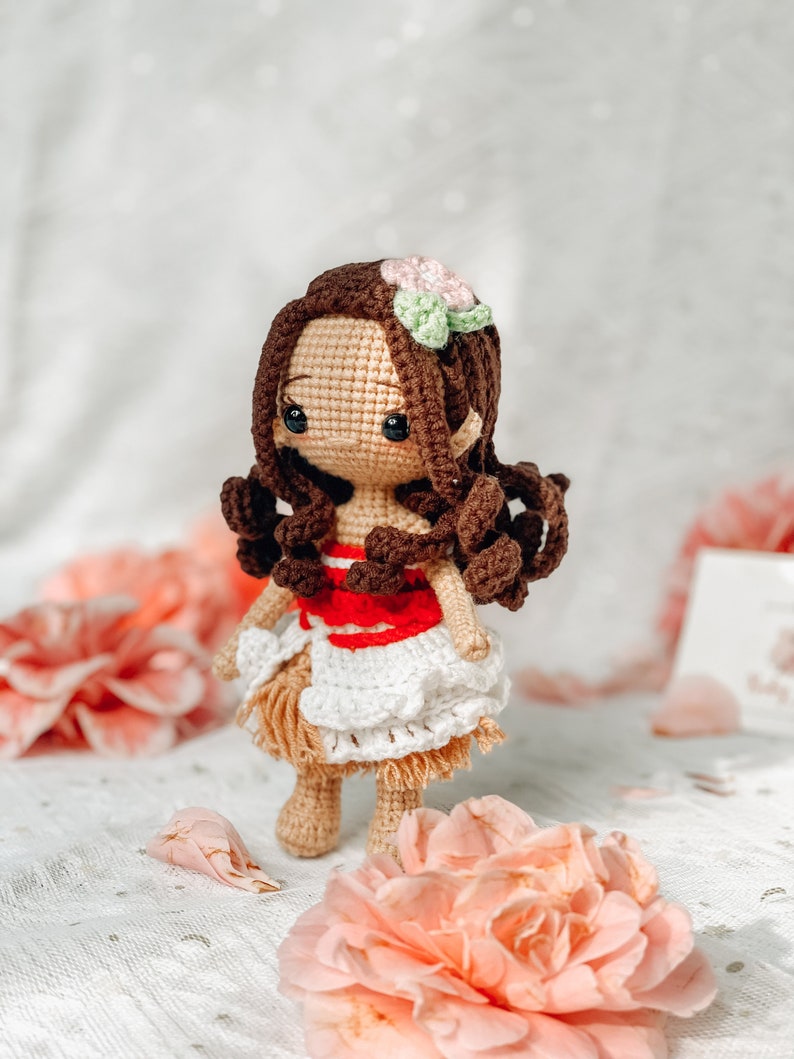May include: A handmade crochet doll with brown hair, a flower hair accessory, and a tan skin tone. The doll wears a red and white top, a white skirt, and a tan skirt. The doll is surrounded by pink flowers.