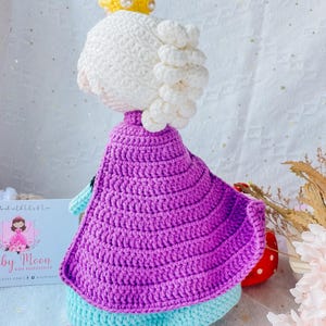 May include: A crocheted doll wearing a purple cape, a white crown, and a light blue dress. The doll has white yarn hair.
