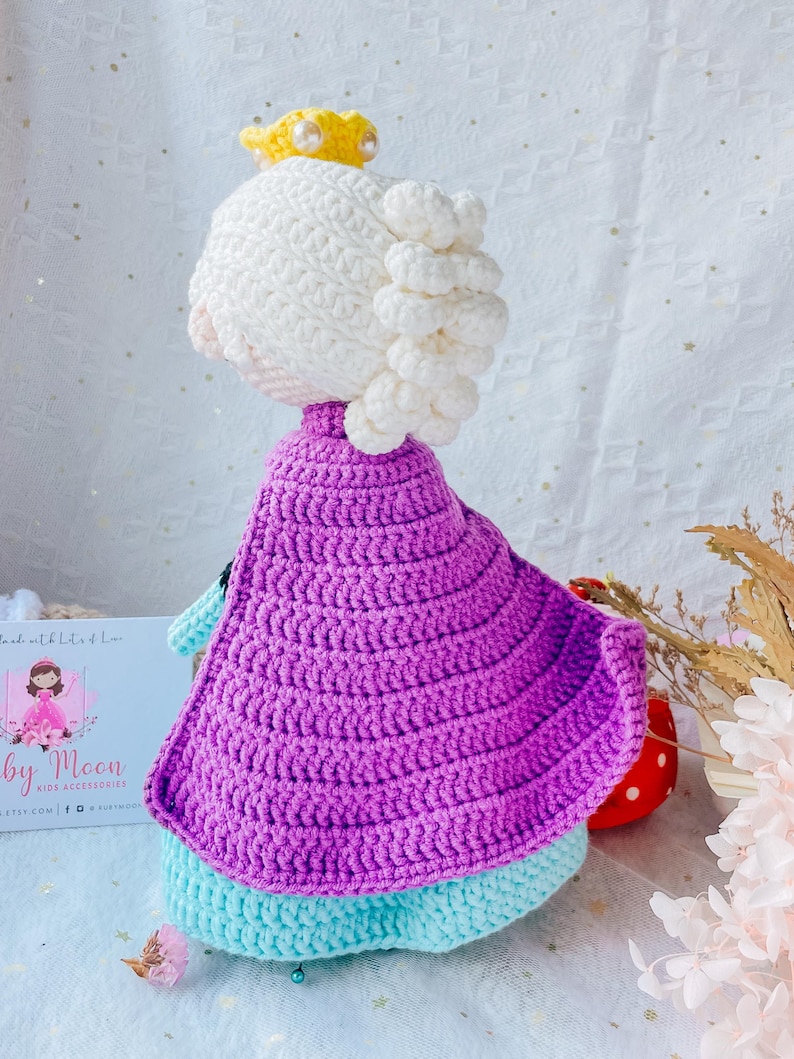 May include: A crocheted doll wearing a purple cape, a white crown, and a light blue dress. The doll has white yarn hair.
