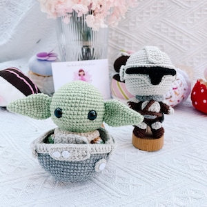 May include: Handmade crochet figures of a green alien in a gray and blue carrier and a Stormtrooper. The alien has large ears and black eyes. The Stormtrooper is gray and brown, standing on a wooden base. A bouquet of pink flowers is in the background.