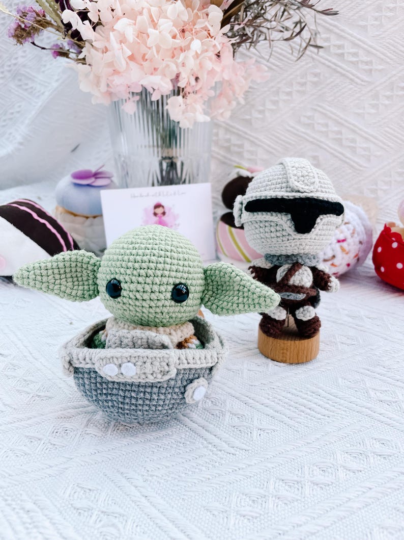 May include: Handmade crochet figures of a green alien in a gray and blue carrier and a Stormtrooper. The alien has large ears and black eyes. The Stormtrooper is gray and brown, standing on a wooden base. A bouquet of pink flowers is in the background.