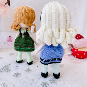 Puede incluir: Dos mu&ntilde;ecas de crochet con el pelo largo, una con el pelo marr&oacute;n y un vestido verde, la otra con el pelo blanco y un vestido azul. Las mu&ntilde;ecas est&aacute;n de pie sobre una superficie blanca con copos de nieve plateados.