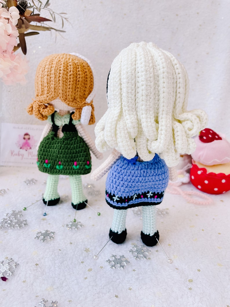 Puede incluir: Dos mu&ntilde;ecas de crochet con el pelo largo, una con el pelo marr&oacute;n y un vestido verde, la otra con el pelo blanco y un vestido azul. Las mu&ntilde;ecas est&aacute;n de pie sobre una superficie blanca con copos de nieve plateados.