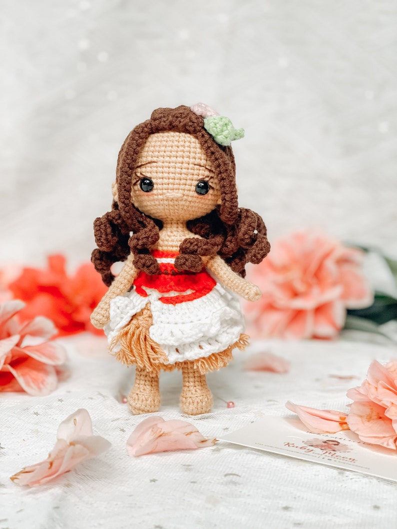 May include: A handmade crochet doll with brown hair, dark eyes, and a floral hair accessory. The doll wears a red and white top and a white skirt with fringe. The doll is standing on a white surface with pink flowers in the background.