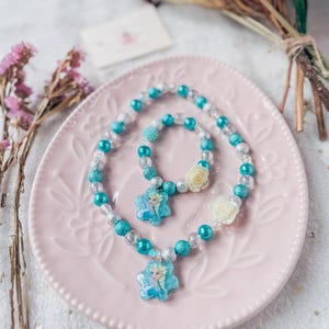 Pode incluir: Um conjunto de colar e pulseira de contas turquesa e prata com um pingente de floco de neve com uma imagem de Elsa de Frozen.