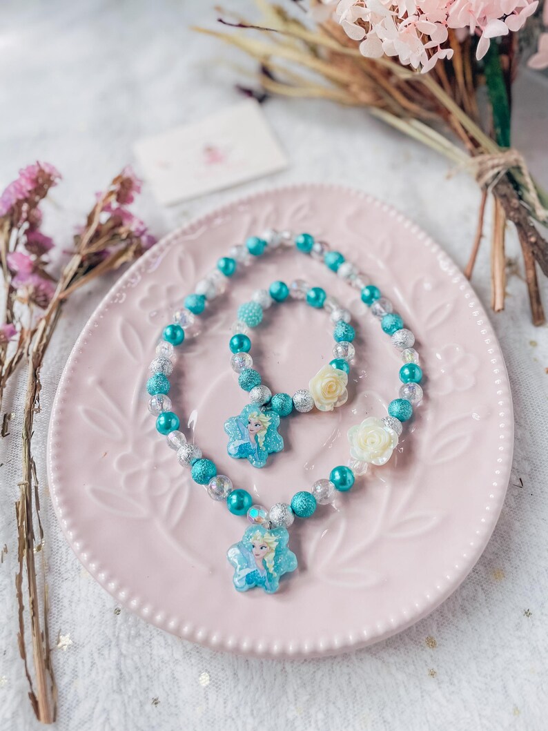 Pode incluir: Um conjunto de colar e pulseira de contas turquesa e prata com um pingente de floco de neve com uma imagem de Elsa de Frozen.
