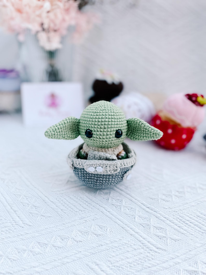 May include: A crocheted figure of a character with large ears and black eyes, seated in a gray and beige crocheted pod. The figure is light green, with a beige body and large, pointed ears. The pod is gray with white accents.