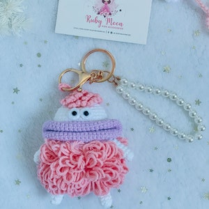 Puede incluir: Un llavero de crochet rosa y blanco con una bufanda morada y ojos grandes. El llavero est&aacute; unido a un llavero de oro y una cadena de perlas.