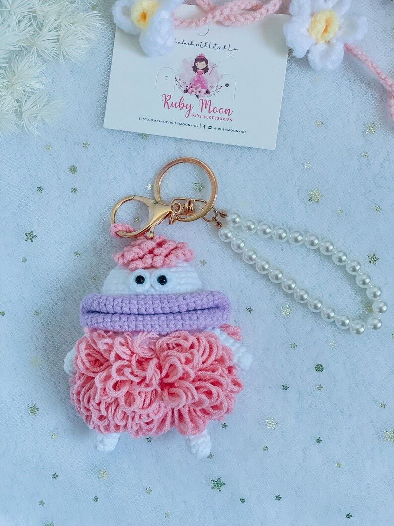 Puede incluir: Un llavero de crochet rosa y blanco con una bufanda morada y ojos grandes. El llavero est&aacute; unido a un llavero de oro y una cadena de perlas.