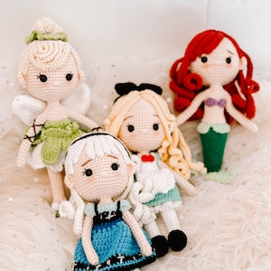May include: Four crocheted dolls, each with a different design and color scheme. The dolls are sitting on a white fluffy surface. The dolls are dressed in various outfits, including a green dress with wings, a blue dress, a white dress with a red heart, and a mermaid tail.