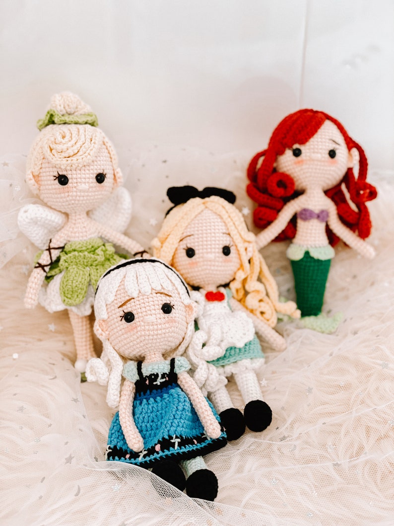 May include: Four crocheted dolls, each with a different design and color scheme. The dolls are sitting on a white fluffy surface. The dolls are dressed in various outfits, including a green dress with wings, a blue dress, a white dress with a red heart, and a mermaid tail.
