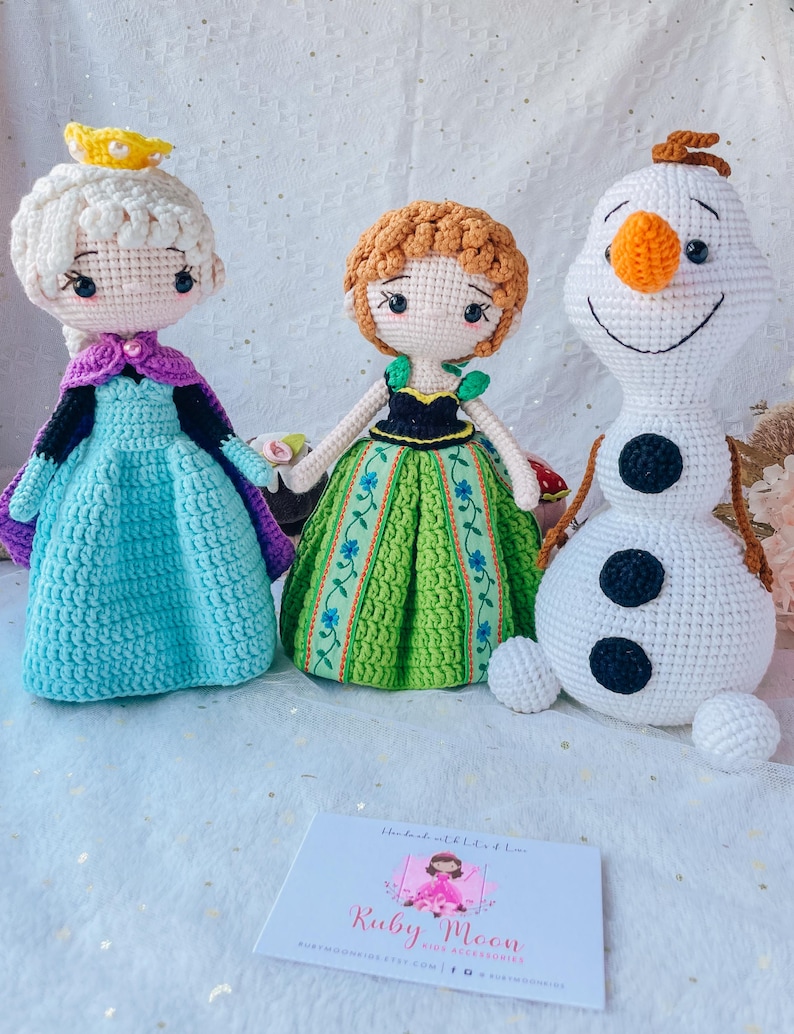 May include: Three handmade crochet dolls: Elsa in a blue dress and purple cape, Anna in a green dress, and Olaf the snowman. The dolls are on a white surface. A business card for Ruby Moon is in the foreground.