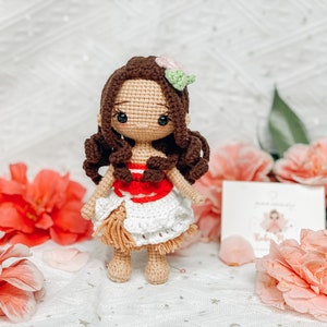 May include: A handmade crochet doll with brown hair and a white and red dress. The doll has a flower accessory in its hair and is standing in front of pink flowers. A small card is visible in the background.