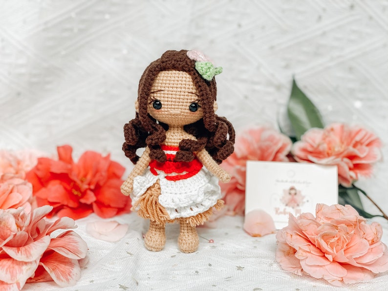 May include: A handmade crochet doll with brown hair and a white and red dress. The doll has a flower accessory in its hair and is standing in front of pink flowers. A small card is visible in the background.